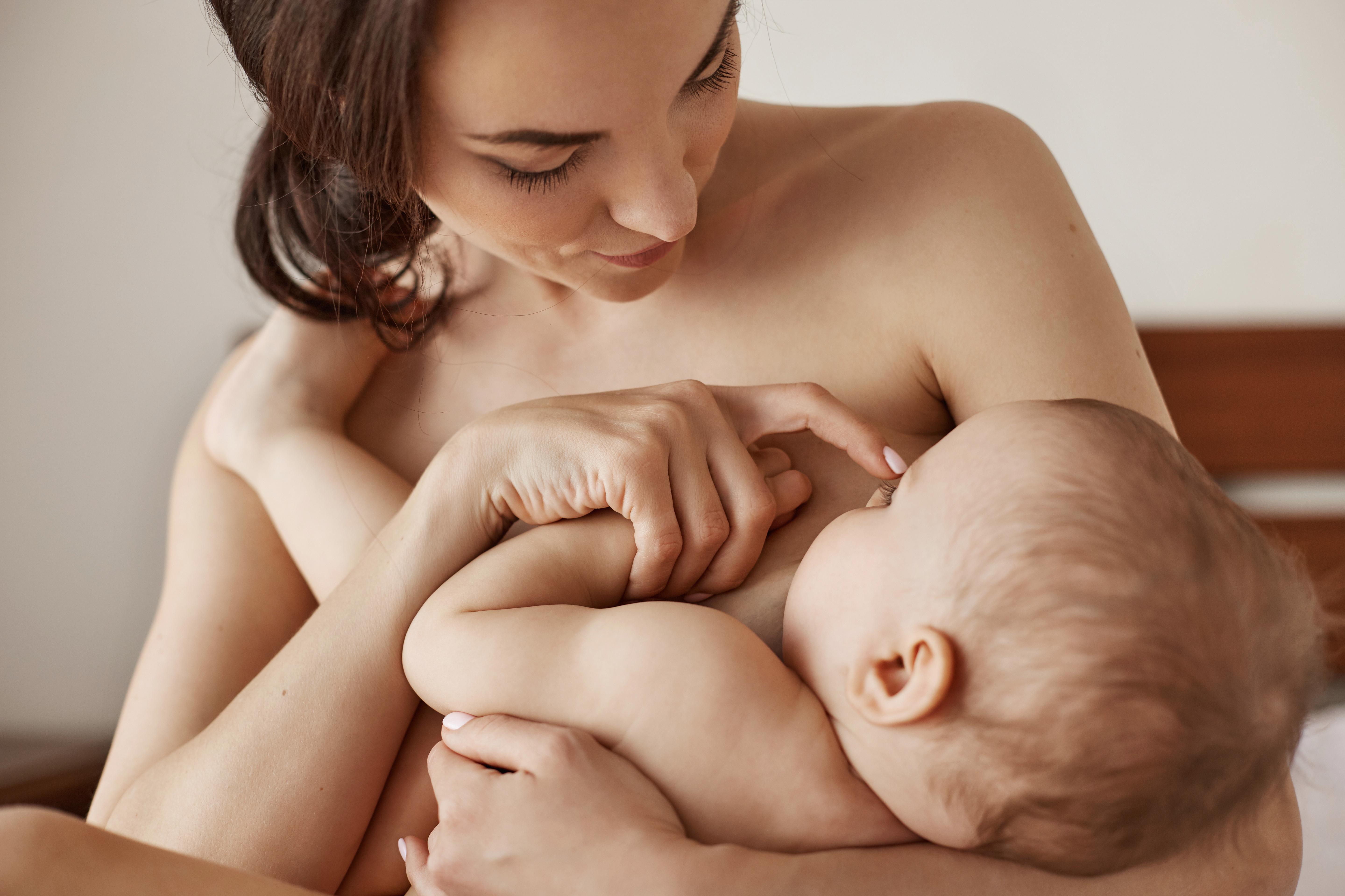 Young tender nude mother breastfeeding hugging her newborn baby sitting in bed at morning.